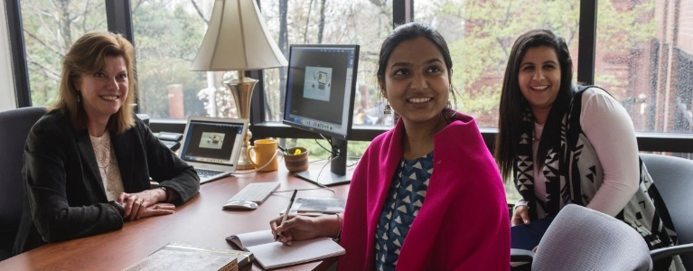 Three women sit at a desk in front of a window