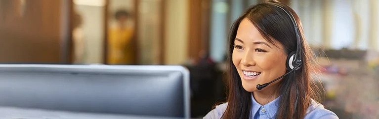 A woman on a headset smiles as she looks at a desktop computer screen.