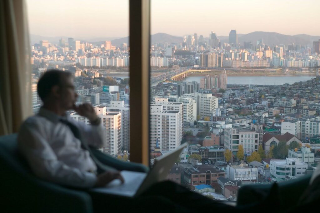 A business person working on a laptop computer looks out an office window revealing the skyline of Seoul, South Korea