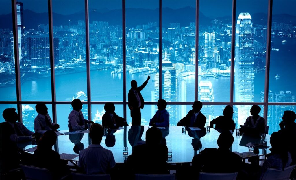 A group of business people in silhouette meet in a conference room; the Hong Kong skyline is visible behind them through a large window.