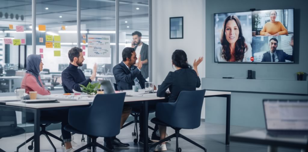 Four working professionals sit at a conference table in an office conference room and address three colleagues virtually on a large screen on the wall.