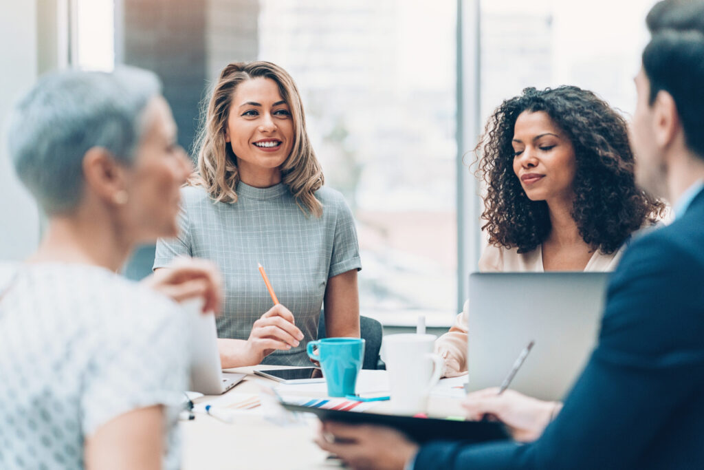 Four professionals speak to each other around a table with laptops, pencils, and a coffee mug.